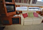 Cozy boat cabin living area with a beige leather sofa and colorful cushions, wood built‑in entertainment center housing a TV and controls, windows with blinds in the background.