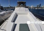 Wide view of a white yacht bow with built‑in seating, docked at a marina on a sunny day, city skyline in the background. The caption reads: 'Bow is made to entertain or just relax at the island!'