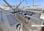 Boat cockpit at a sunny marina: steering wheel, Garmin display, throttle levers, bench seat, and a view of moored yachts through the windshield.