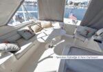 Spacious white yacht flybridge seating area with a central table and cushions, marina seen through windows; flag visible outside.