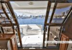 Open sliding glass doors reveal a sunlit yacht cockpit with white seating, looking out over a marina full of boats.