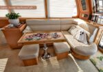 Cream U-shaped yacht lounge with a wooden table and floral runner, two small stools, and a potted plant by the window blinds.