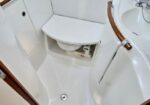 Compact white boat bathroom with a central toilet, curved wooden handrails, and a sink on the right; door open.