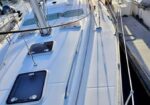 View along the deck of a white sailboat docked at a marina, with masts and other boats in the background at sunset.
