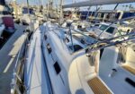 Close-up view along the deck of a white sailboat at a crowded marina, with masts, rigging, and neighboring boats on both sides.