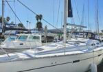 White sailboat docked at a marina with other boats, masts, and palm trees visible on a sunny day.
