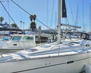 White sailboat docked at a marina with other boats, masts, and palm trees visible on a sunny day.