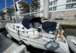 White Beneteau 323 sailboat docked at a marina, with blue cockpit covers, fenders, and modern buildings in the background.