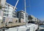 White sailboat moored at a marina beside modern apartment buildings under a clear blue sky.