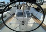 Sailboat cockpit with a large stainless steering wheel, navigation display, and wood-decked floor, marina in the background.