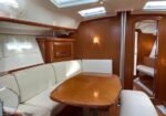 Interior of a yacht cabin with a wooden oval table and cream leather seating along the walls, warm lighting, and a doorway to another cabin.