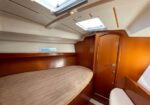 Boat cabin with a single bed along the left, warm wood walls, built-in shelf, and a closed wooden door nearby under overhead skylights.