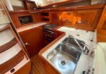 Compact boat galley with polished wooden cabinets, a stainless double sink, a two-burner stove, built-in microwave, and curved ladder steps nearby.