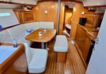 Cozy sailboat cabin with a U-shaped white-cushioned dinette around a teak table; cups sit on the table and wood-paneled walls surround the space.