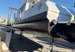 White passenger boat raised on a trailer in a boatyard, supported by stands with equipment and shadows on the concrete ground