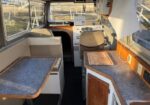 Interior of a small boat cockpit with a captain's chair, helm/navigation area, and a blue-counter galley on the left side.