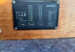 Inverter/charger control panel labeled Xantrex mounted on a wooden wall, showing DC volts and amps scales and buttons: Setup, Charge, Invert, Power Share