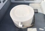 White cylindrical portable toilet on a boat deck with a gray hose attached to the side, secured on a flat, light-gray surface.
