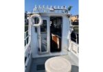 White boat deck with an open cabin door, a life preserver hanging on the left, and a round table in the foreground under a clear blue sky.