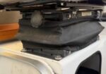 Black padded cargo bag strapped to a roof rack on a white vehicle, secured with metal clamps for extra luggage storage.