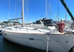 White sailboat moored in a marina with tall masts, blue sky, and a coiled red rope on the bow.
