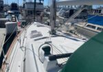 On the deck of a white sailboat in a marina, with lines, winches, and fittings, and other boats moored nearby