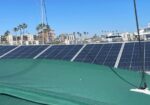 Blue solar panels cover a green rooftop with a marina and palm trees in the background.