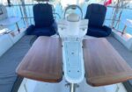 Catamaran cockpit with two blue captain's chairs, teak folding tables extended over bench seating, and a central steering console by the marina.