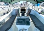 Sailboat cockpit with central steering wheel, Raymarine navigation display, and long cushioned benches on both sides at a marina.