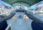 Inside a white sailboat cockpit at a marina, showing the helm console with the steering wheel, green canopy, and blue cushioned seating; hull number 1200253 visible.
