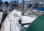 Top-down view of a white sailboat deck in a marina, with rigging, winches, hatches, and neighboring boats docked nearby.
