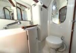 Compact white yacht head with sink, mirror, and toilet; wooden trim and rounded porthole window.