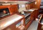 Compact sailboat galley with wood cabinetry, microwave on left, gas stove with a kettle, and a sink with curved faucet, netting above and striped deck flooring