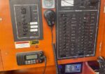 Close-up of a boat's electrical control panel with labeled breakers, a handheld VHF radio, and a digital display.