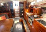 Interior of a sailboat cabin with glossy wood cabinetry, a compact galley kitchen, stove, sink, and seating area around a wooden table amid warm natural light.