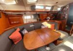 Wood-paneled yacht salon with a curved gray sofa around a teak table, orange cushions, and the navigation/navigation area visible in the background.