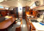 Sunlit wooden sailboat interior with a dining table and bench seating, a galley on the right with a sink and kettle, and stairs to the cabin ahead.