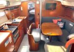 Sailboat cabin with a wooden galley on the left (sink, stove, oven) and a U-shaped dinette on the right; doorway to a small forward cabin and a wall-mounted TV.