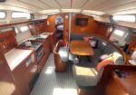 Interior of a sailboat: compact galley with stove and sink, U-shaped seating around a wooden table, TV on wall, and a doorway to a sleeping cabin.