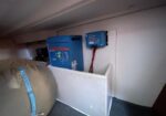 Blue marine inverter/charger mounted on a white bulkhead in a boat cabin, connected with red and black cables; a beige life raft sits to the left.