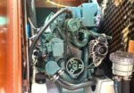 Marine diesel engine in a small engine room: teal-green block with belts, hoses, and an alternator visible, mounted on a white base.