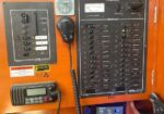 Boat electrical control panel with numerous labeled circuit switches, a handheld mic, a digital voltmeter, and a 12V power board on a wood dashboard