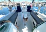 Cockpit of a sailboat with long blue-cushioned bench seats along both sides, central helm area with two blue captain chairs, a small table, and marinas and other boats in the background.
