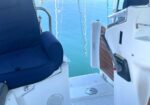 Blue cushioned seat on a white boat cockpit with calm blue water in the background, near a wooden hatch and railings.