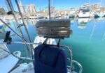 Barbecue grill mounted on the stern rail of a sailboat at a marina with calm blue water and city buildings in the background, blue seating nearby.