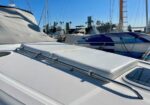 White boat deck with a raised hatch and stainless railing, at a sunny marina with other boats in the background