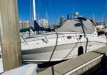 White motor yacht moored at a marina with a gray cover, dock pilings and a city skyline under a blue sky.