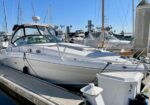 White motor yacht docked at a marina with other boats and tall masts in the background.