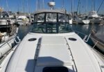 White motor yacht at a marina, bow deck with hatch, cockpit enclosed by windshield and canvas cover, moored among sailboats on calm water.