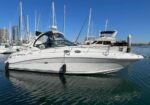 White motor yacht docked at a marina with sailboats and a city skyline in the background on a sunny day.